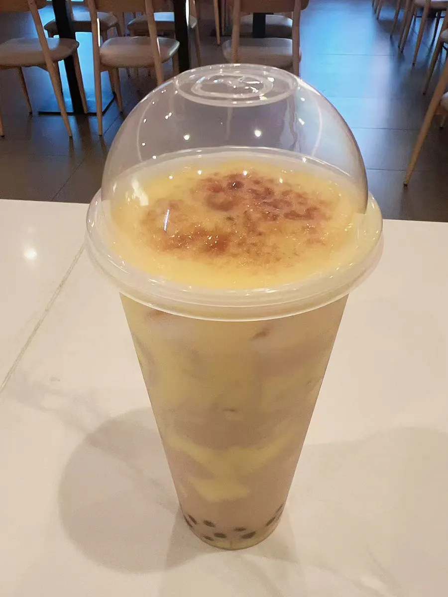 Brulee top creamy milk tea tapioca pearls clear dome plastic cup white table at Mochi & Crunch Treats, a Modern Dessert Café in Tupelo